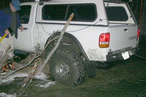 2003 Jeep TJ - A Ford pickup truck appears to be stuck in a muddy, watery area, with a person holding an axe standing next to it.