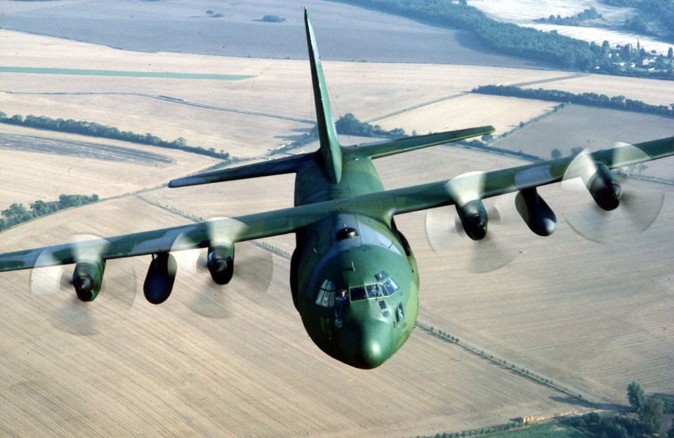 Green camouflaged four-engine military turboprop transport plane flying over fields