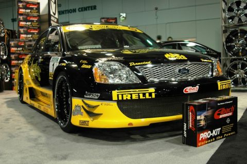 1991 Ford Taurus - A modified black and yellow sedan with numerous sponsor decals is displayed indoors.