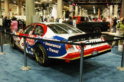 1991 Ford Taurus - A race car with a red, white, and blue livery is displayed indoors with a black pickup truck in the background.