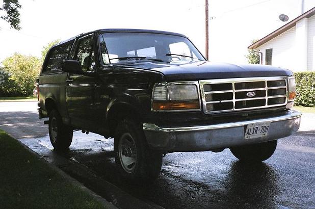 Black Ford Bronco SUV parked curbside on a wet street, front three-quarter view with chrome grille.
