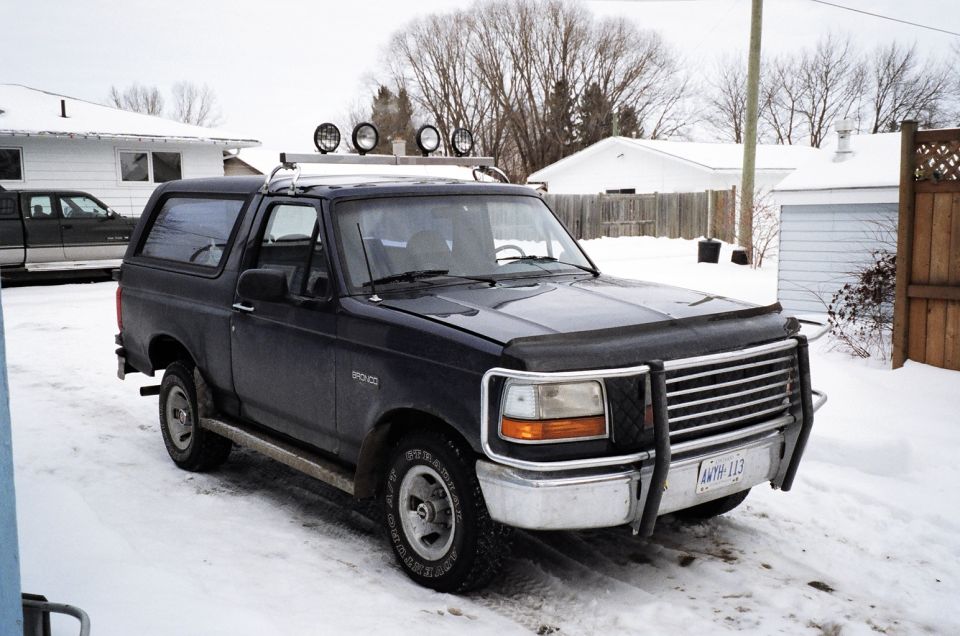 Dark blue Ford Bronco with a brush guard and roof lights parked in the snow. Another truck is visible behind it