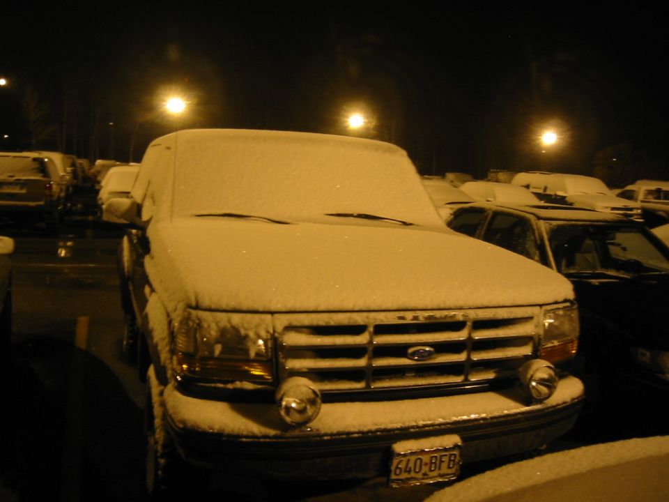 Snow-covered Ford truck with round bumper driving lights in a lot at night