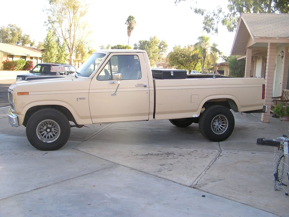 Tan Ford F-150 regular cab long-bed pickup, driver-side view, black bed toolbox, parked in driveway