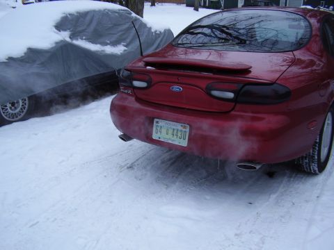 1997 Ford Taurus - A red sedan with its exhaust steaming sits in the snow next to another vehicle covered by a tarp.