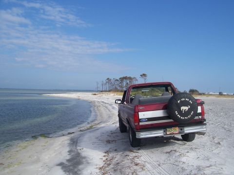 1995 Ford Bronco - Rear three-quarter view of a red Ford Bronco with its hardtop removed, parked on a white sand shoreline with calm water and a stand of pine…
