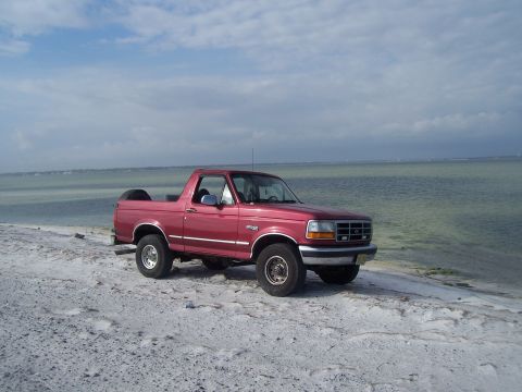 1995 Ford Bronco - A red 1995 Ford Bronco is parked on a sandy beach with the ocean in the background.