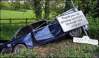 1972 Ford Ranchero - A car has crashed off the road and is leaning against a sign that reads 'Thank you for driving carefully through the village'.