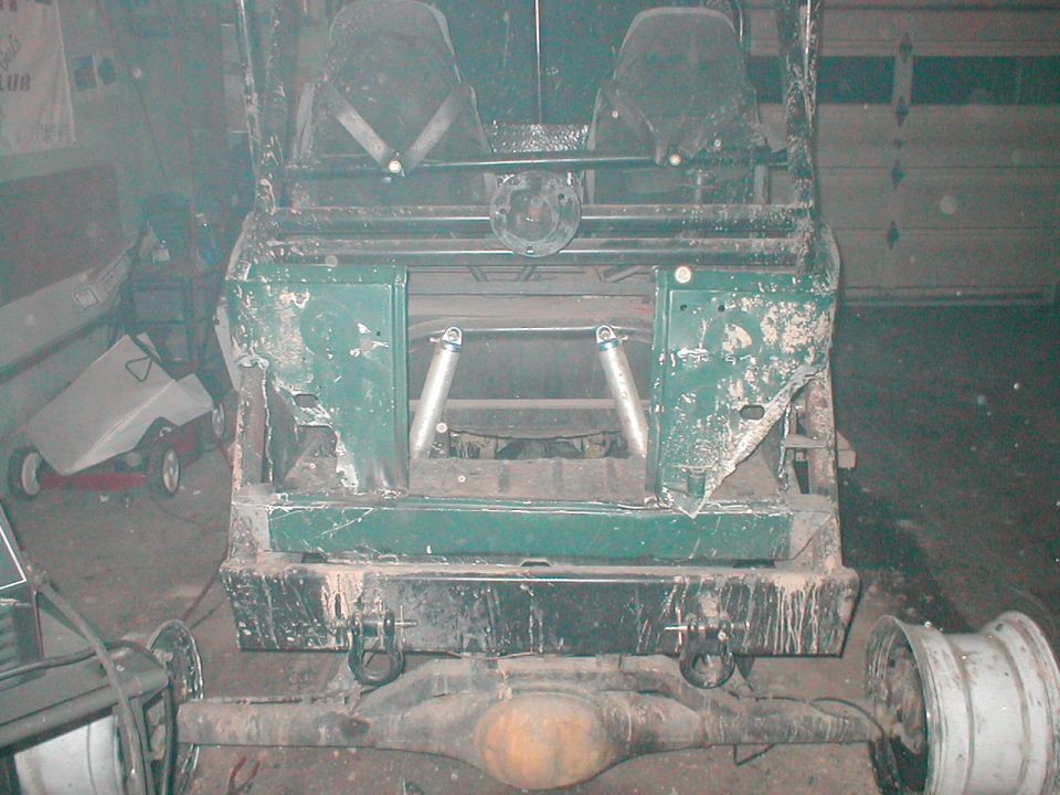 Rear frame and suspension of a green vehicle, possibly a Bronco, with two seats visible. The body is damaged