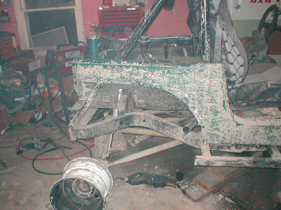 Green off-road vehicle chassis with roll cage, undergoing repairs in a garage. A wheel is detached and lying on the floor