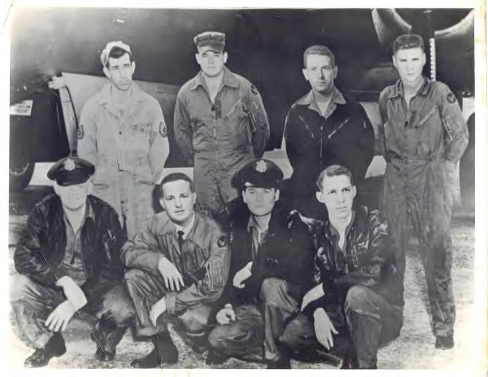 Group of nine men in flight suits and uniforms posing in front of an aircraft