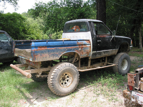 Rusty pickup truck with blue bed, lifted suspension, and large tires, parked outdoors