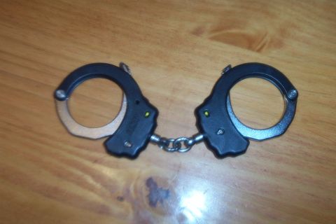 1998 Ford Crown Victoria - A pair of black metal handcuffs with a silver chain are displayed on a wooden table.
