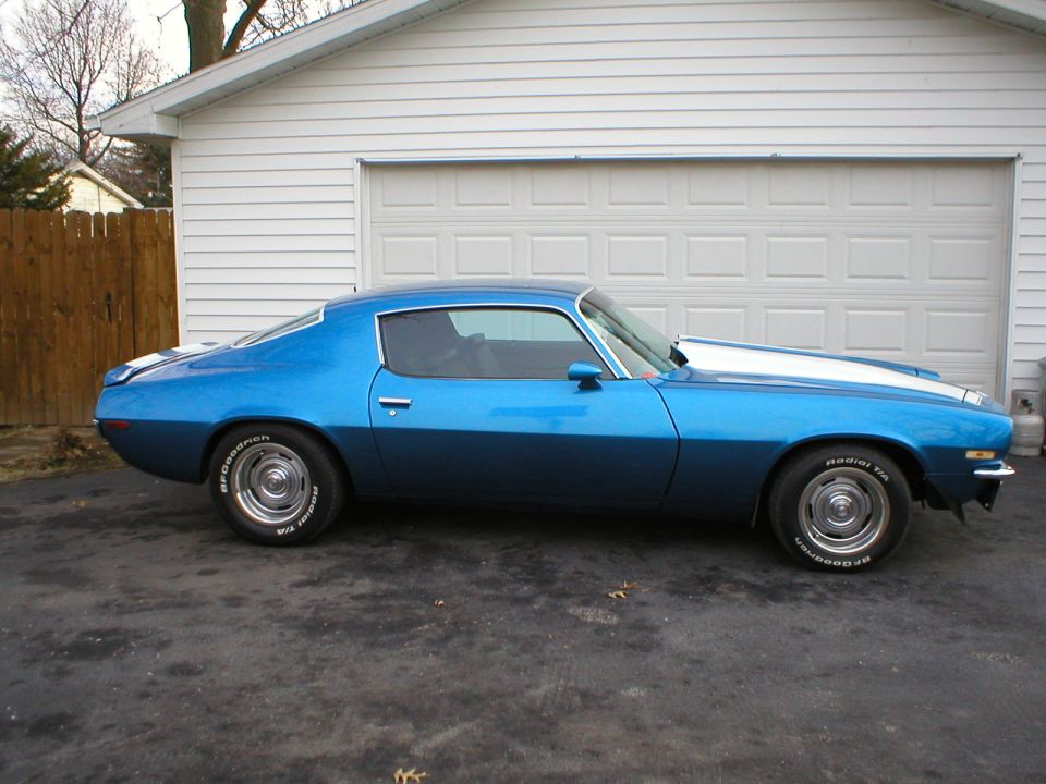 Blue 1970 Chevrolet Camaro parked in front of a garage, side view with white racing stripes on the hood and trunk