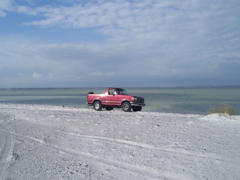 1995 Ford Bronco - A red SUV sits on a sandy shore with the ocean in the background under a cloudy sky.