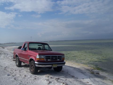 1995 Ford Bronco - A red pickup truck is parked on a sandy beach next to the ocean under a cloudy sky.