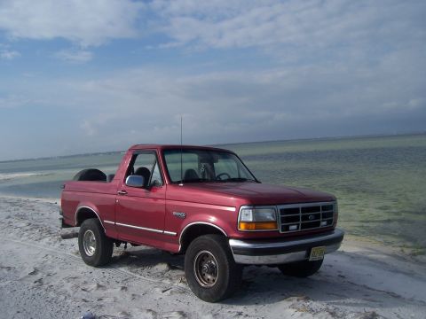1995 Ford Bronco - This red 1995 Ford Bronco is parked on a sandy beach next to the water.