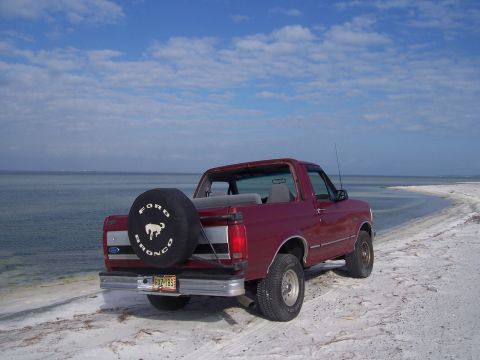 1995 Ford Bronco - Rear three-quarter view of a red Ford Bronco with its hardtop removed, parked on a white sand spit at the water's edge under a partly cloud…