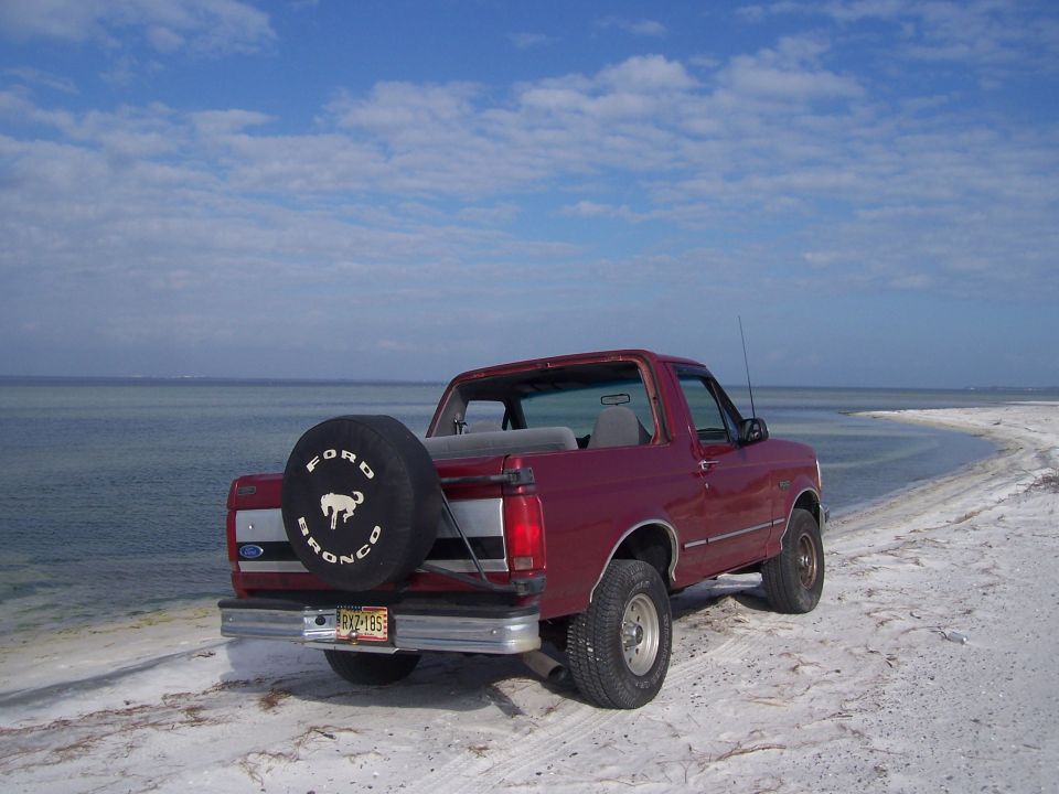 Topless red Ford Bronco on white sand at the shoreline with Bronco spare tire cover