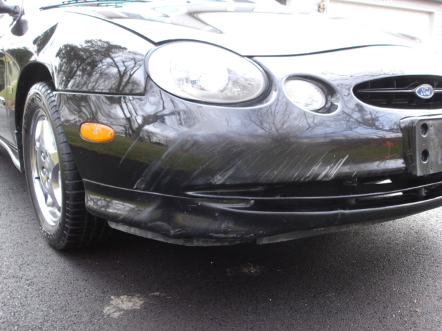 Black 1998 Ford Taurus front bumper with significant scratches and dents