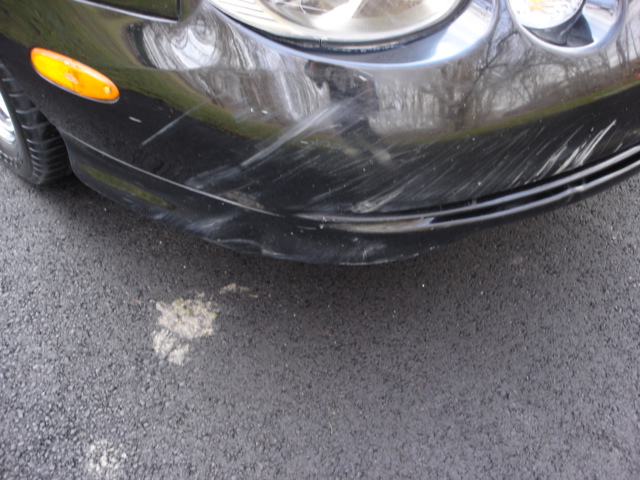 Close-up of a black car's front bumper showing numerous scratches and a damaged lower section. An amber turn signal is visible