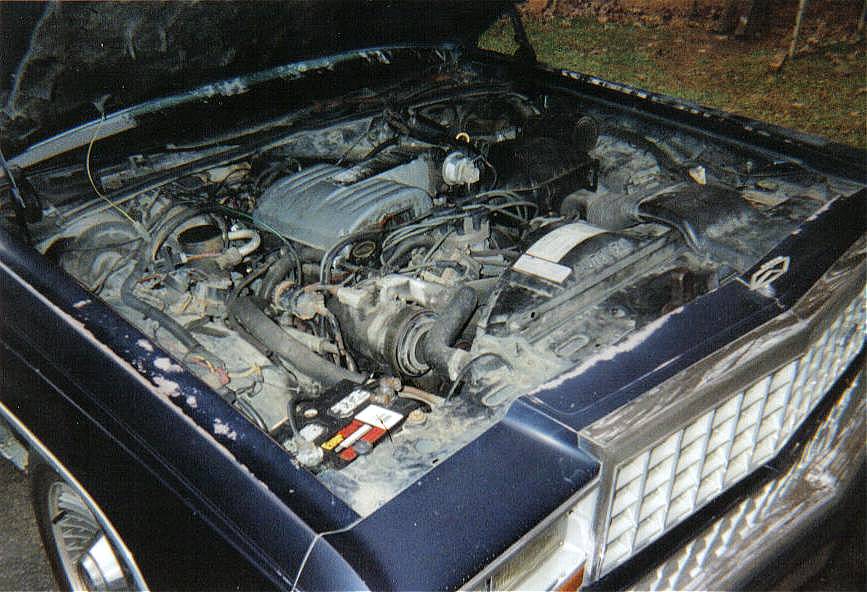 Open engine bay of dark blue sedan showing dusty V-engine and hoses