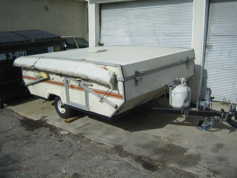 1990 Ford Bronco - collapsed white pop-up tent trailer with an orange accent stripe and propane tank sits on pavement beside a roll-up door.