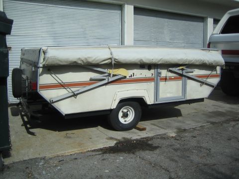 1990 Ford Bronco - folded white pop-up tent camper trailer with an orange stripe and crossed support arms parked beside a building.