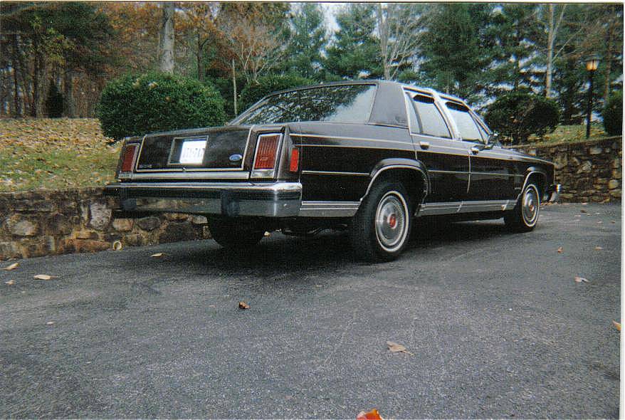 Dark brown Ford LTD Crown Victoria sedan, rear three-quarter view, parked on asphalt by a stone wall