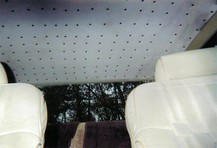 Car interior showing perforated gray headliner, white cloth headrests, and trees through rear window