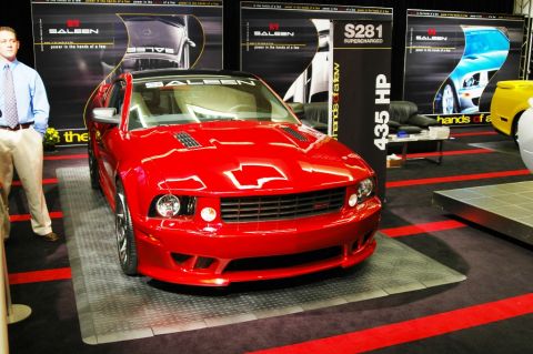 1991 Ford Taurus - A red Saleen S281 Supercharged Mustang is displayed at an auto show with other vehicles and promotional banners.
