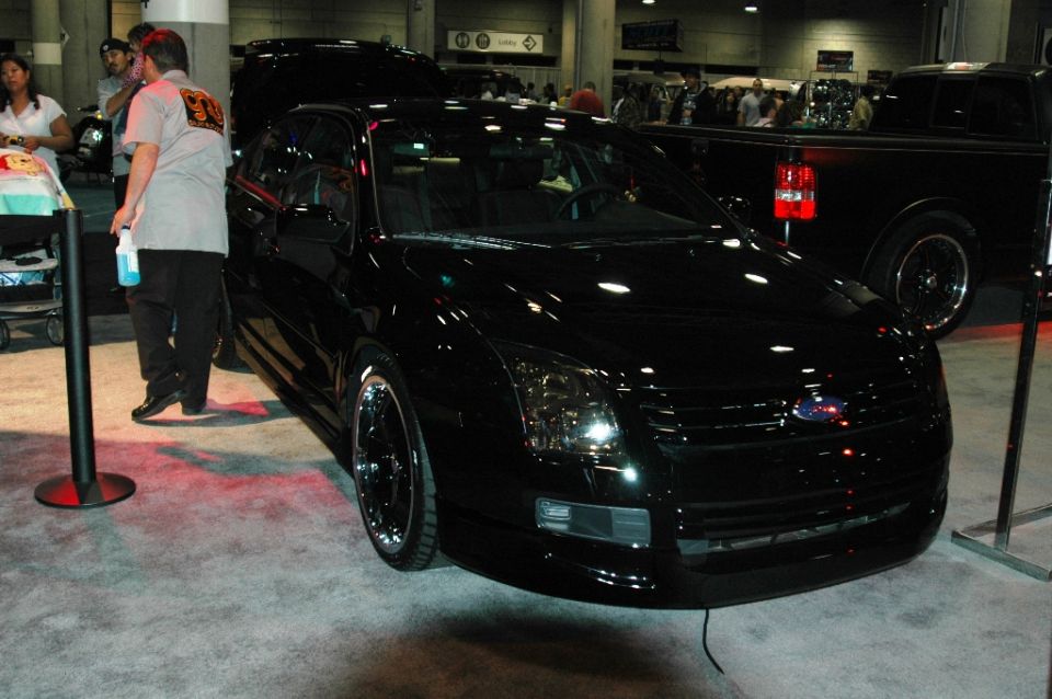 Black Ford Fusion lowered on chrome wheels at an indoor car show, people standing nearby