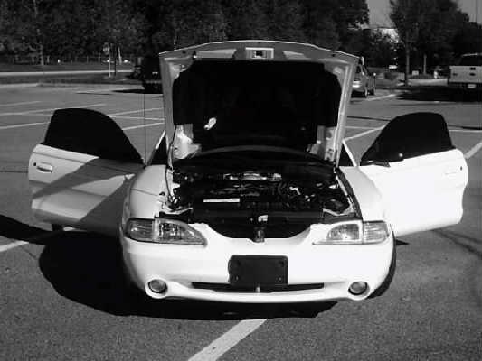 White Mustang coupe from the front with hood raised and both doors open in a parking lot, black-and-white photo