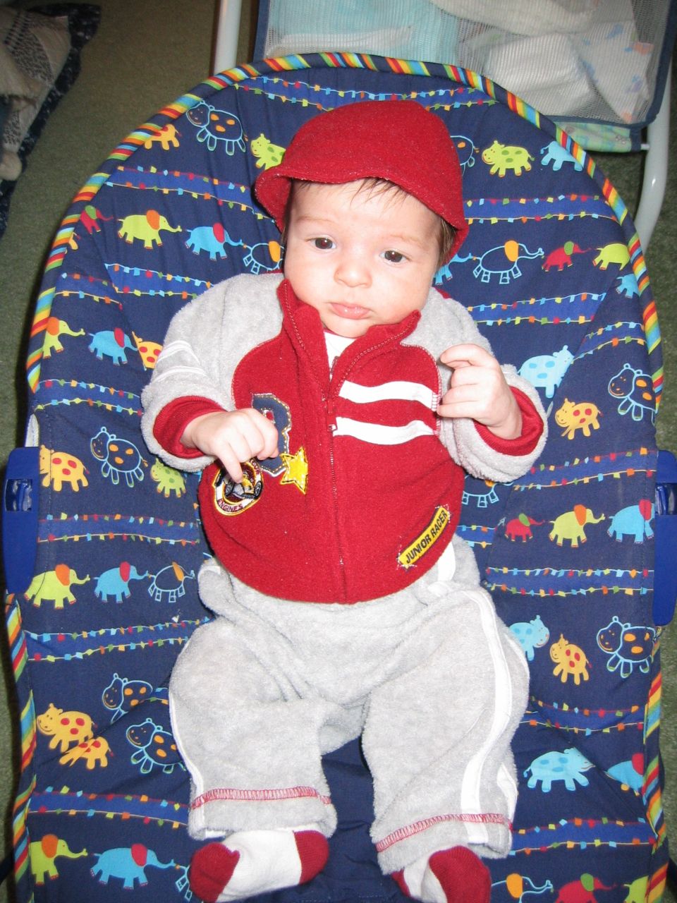 Baby in red cap and Junior Racer track suit sitting in a patterned baby bouncer