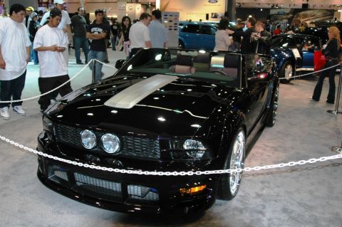 1991 Ford Taurus - A black convertible sports car is displayed at an indoor event with other vehicles and people around.