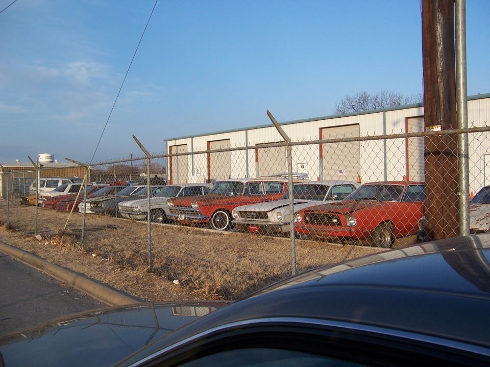 Several cars parked in a lot behind a chain-link fence, with a building in the background