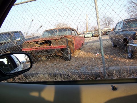 1979 Ford Ranchero - Junkyard scene with a derelict car in the foreground, viewed through a fence.