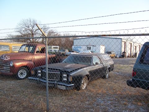 1979 Ford Ranchero - A black 1979 Ford Ranchero sits in a lot behind a fence, next to a rusty pickup truck.
