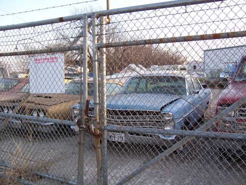 1979 Ford Ranchero - A blue sedan with a distinctive grille is parked behind a fence, with other vehicles visible.