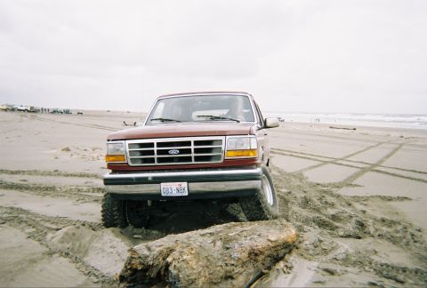 1992 Ford Bronco - A red Ford F-Series truck is stuck on a log on the beach with tire tracks in the sand.