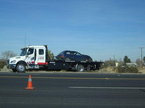1990 Ford Bronco - white flatbed tow truck carries a blue sports car along a two-lane desert road, an orange traffic cone in the foreground.