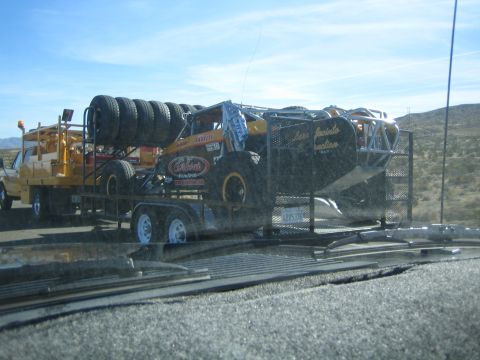1990 Ford Bronco - yellow tube-chassis off-road race buggy with spare tires loaded on a flatbed trailer being towed on a desert highway.
