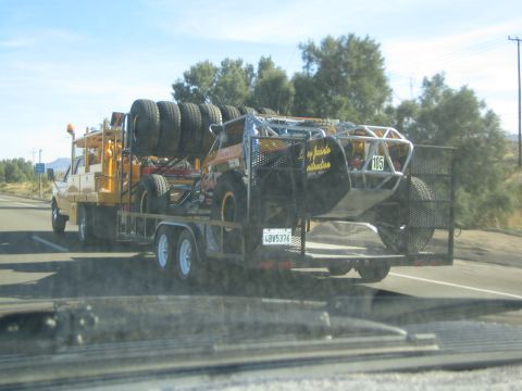 1990 Ford Bronco - yellow off-road race buggy marked 105 with stacked spare tires rides on a flatbed trailer towed along a desert road.