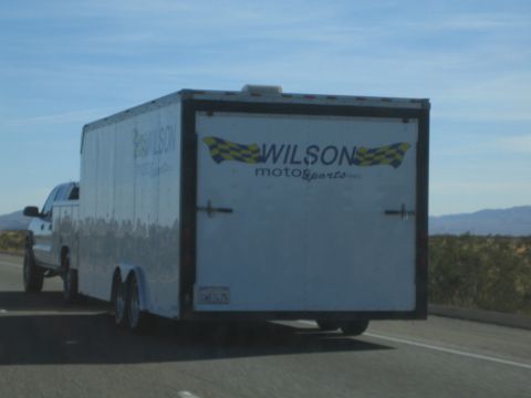1990 Ford Bronco - white tandem-axle enclosed trailer lettered Wilson Motorsports is towed by a pickup along an open desert highway.