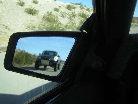 1990 Ford Bronco - vehicle side mirror reflects a lifted dark pickup truck following behind on a desert highway, viewed from inside the cab.