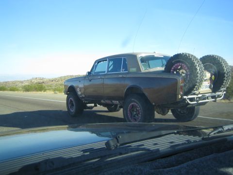 1990 Ford Bronco - brown lifted vintage sedan on oversized tires with two bed-mounted spares puffs exhaust while driving on a desert highway.