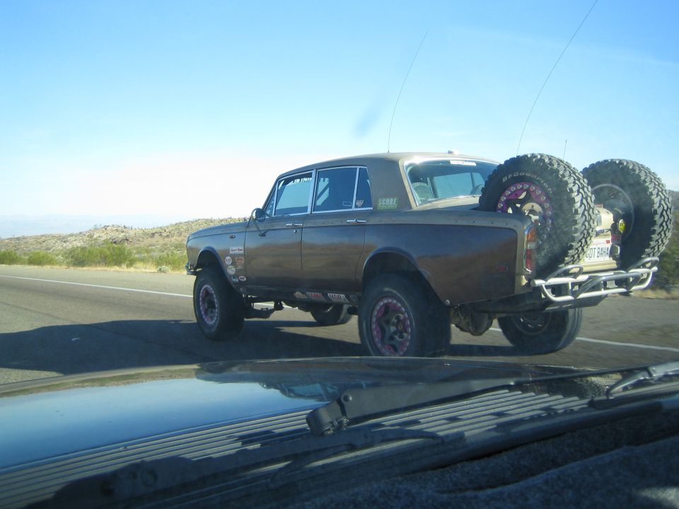Brown lifted vintage sedan with bed-mounted spares emitting exhaust on a highway