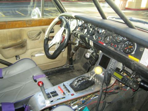 1990 Ford Bronco - stripped interior of a race-prepped car showing a roll cage, aftermarket gauges, radios, switch panel, and a harnessed seat.