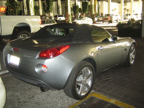 1990 Ford Bronco - Rear three-quarter view of a gray Pontiac Solstice roadster with its black soft top raised, parked in a covered lot.