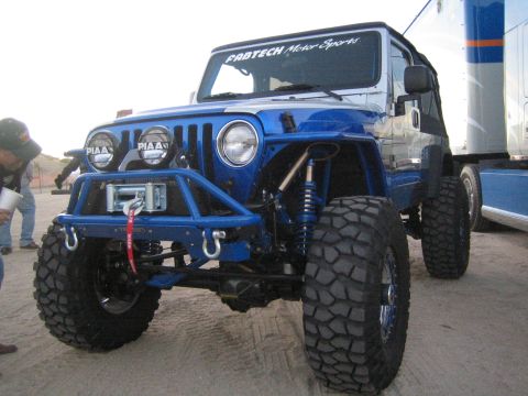 1990 Ford Bronco - blue lifted Jeep Wrangler with a tubular front bumper, winch, PIAA lights, and oversized mud tires beside a trailer.